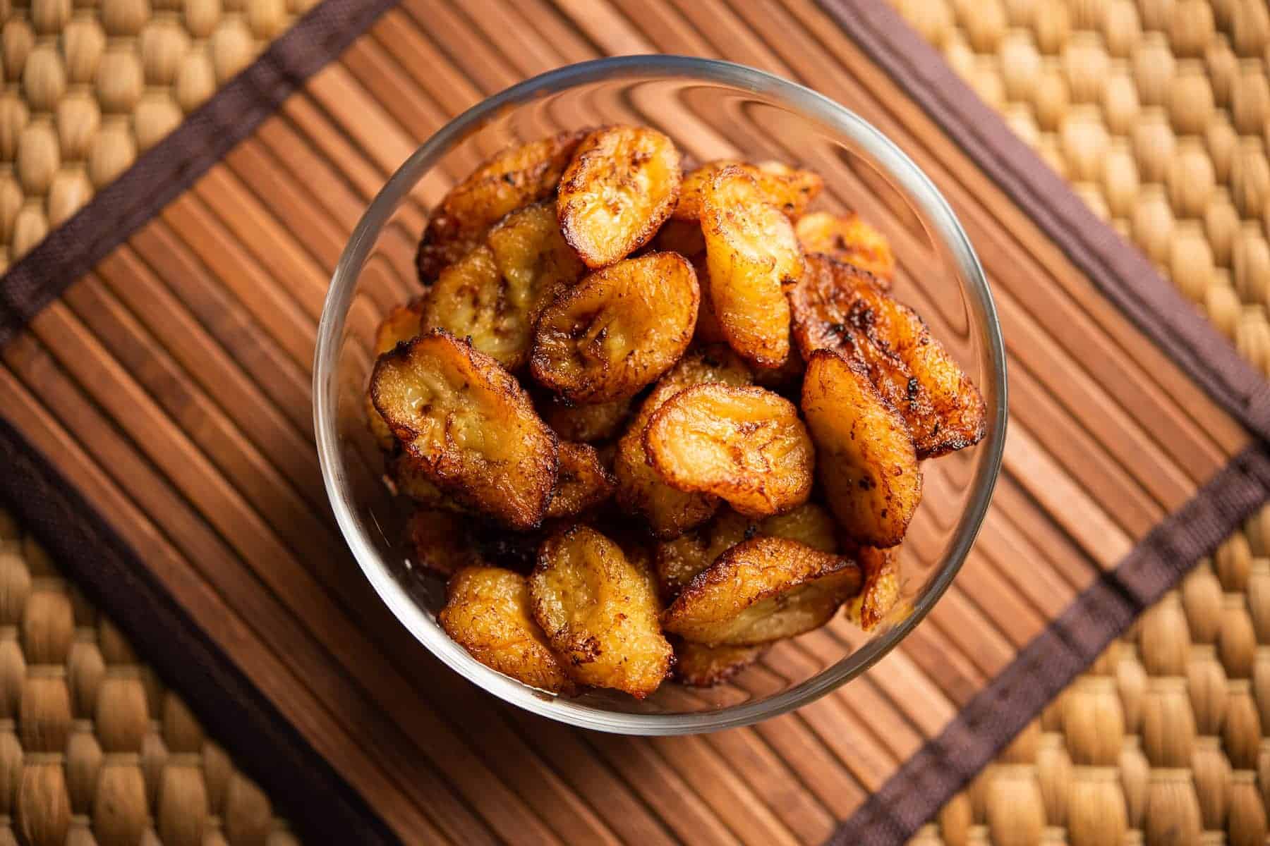 A glass bowl filled with fried plantain slices, a popular choice among Bahamas breakfast foods, sits on a wooden placemat with a woven background.