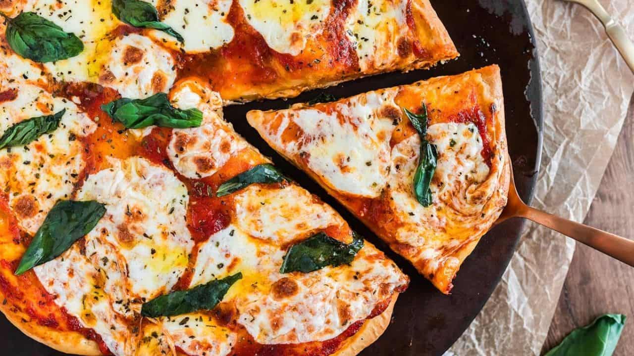 A Margherita pizza with melted mozzarella, tomato sauce, and basil leaves, with one slice being lifted from the whole pizza.