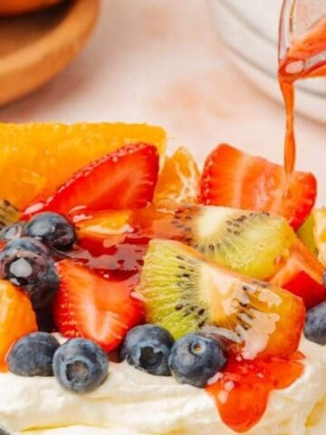 A pavlova topped with whipped cream, sliced kiwi, strawberries, blueberries, orange segments, and fruit sauce being poured over the top.