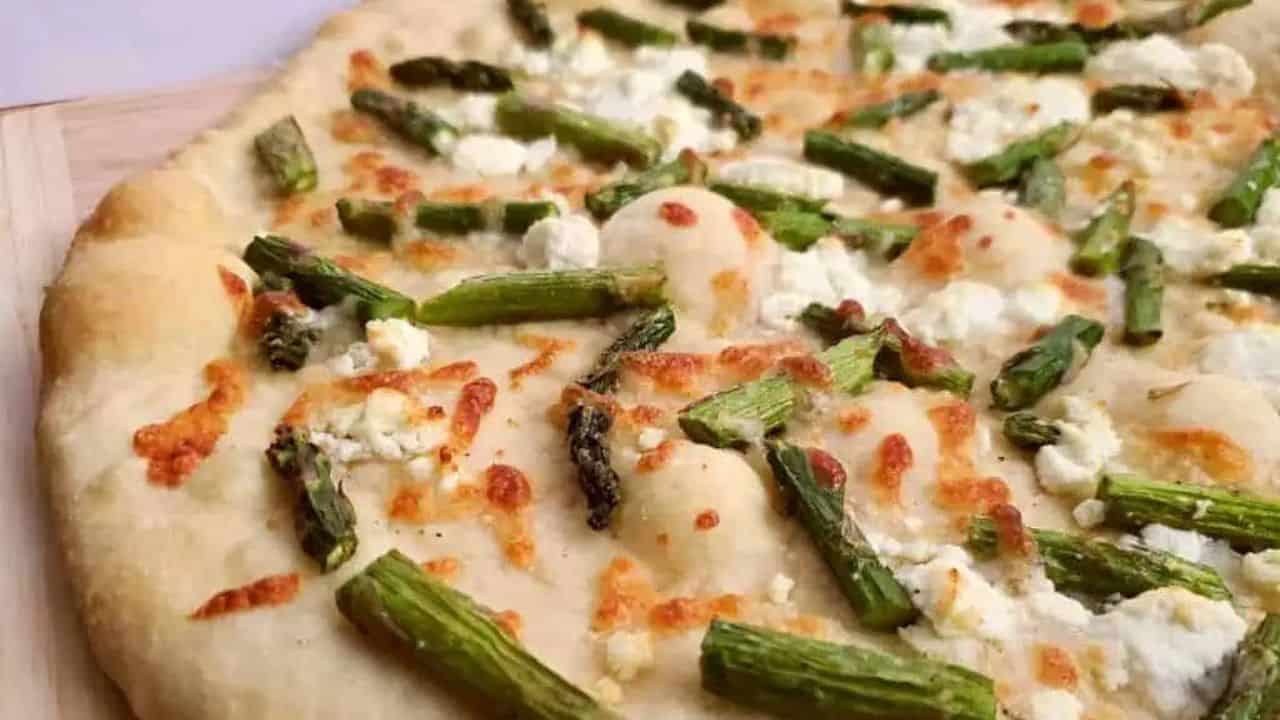 Image shows a close up of an Asparagus and Goat Cheese Pizza on a pizza peel sitting on a black counter.