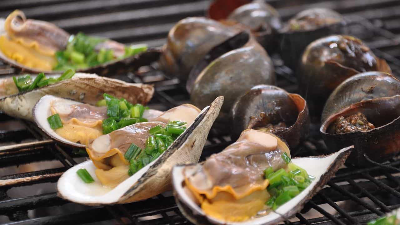 Assorted shellfish, including clams and snails, being grilled on an open flame with chopped green onions as garnish.