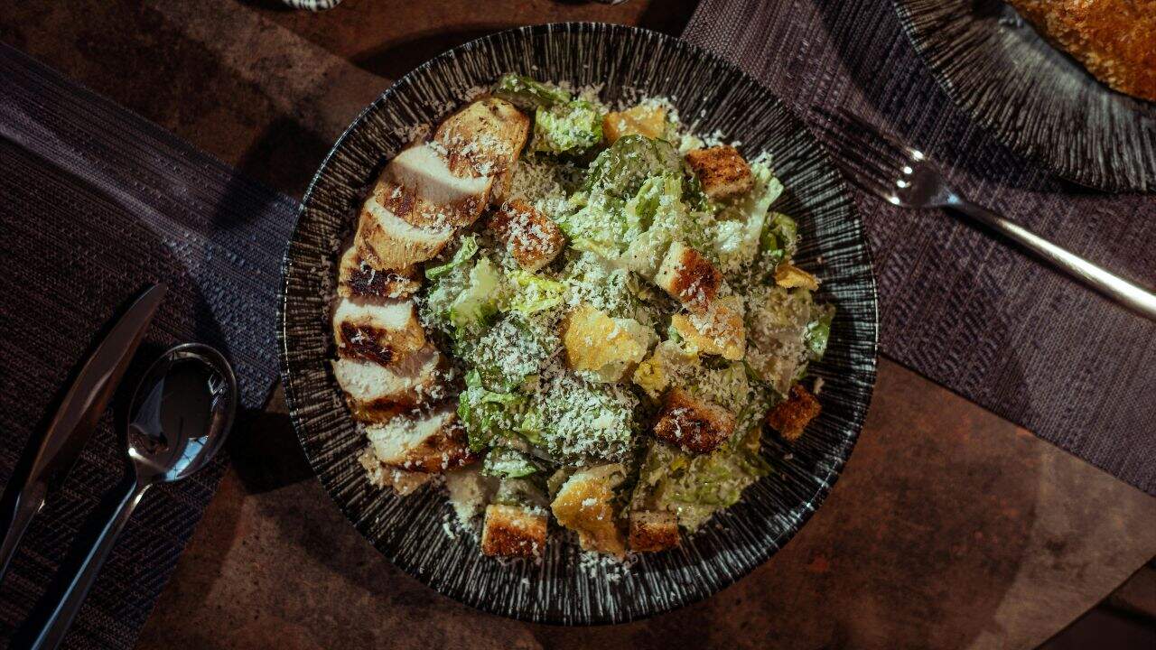 A black bowl filled with Caesar salad topped with grilled chicken slices, croutons, and grated cheese, placed on a set dining table.
