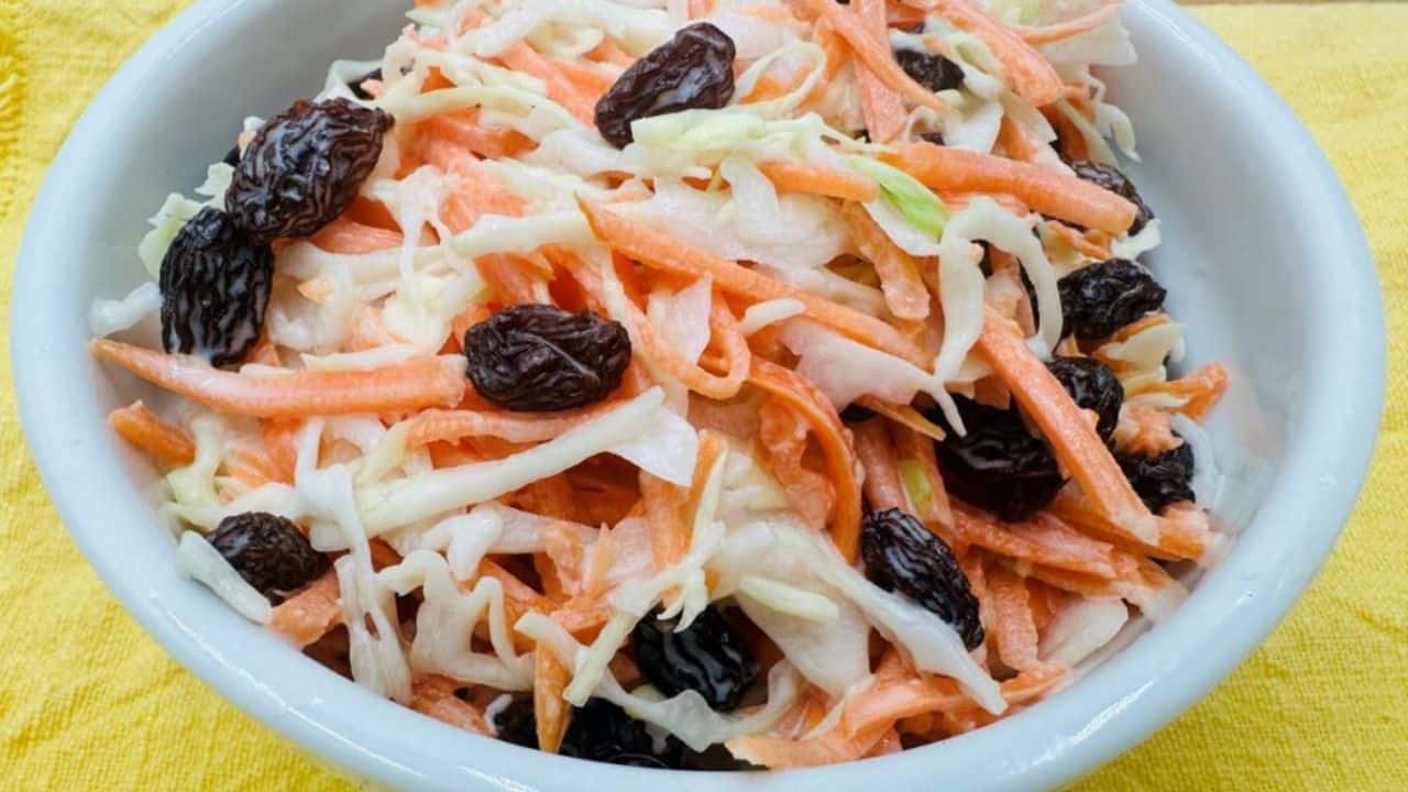 A bowl of coleslaw with shredded carrots and raisins on a wooden surface, next to a red and white checkered cloth.