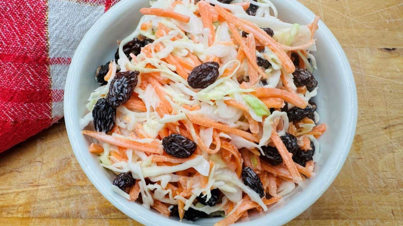 A bowl of coleslaw with shredded carrots and raisins on a wooden surface, next to a red and white checkered cloth.