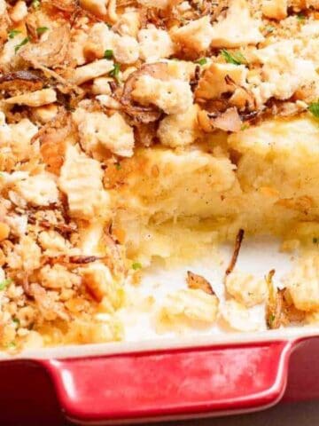 A red baking dish holds a partially served casserole topped with crispy onions and parsley, revealing a creamy filling beneath.