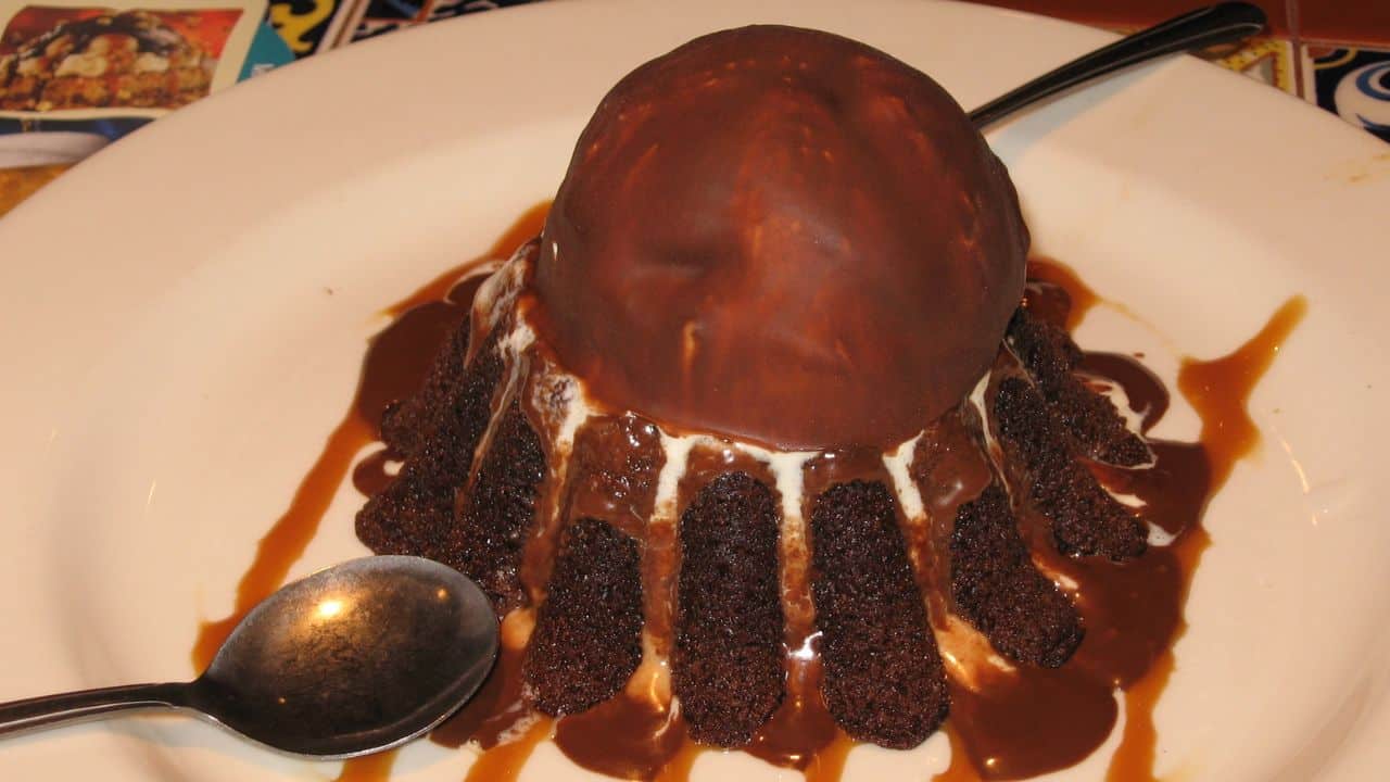 A chocolate dome dessert with cake segments and ice cream, drizzled with caramel sauce, on a white plate with a spoon beside it.