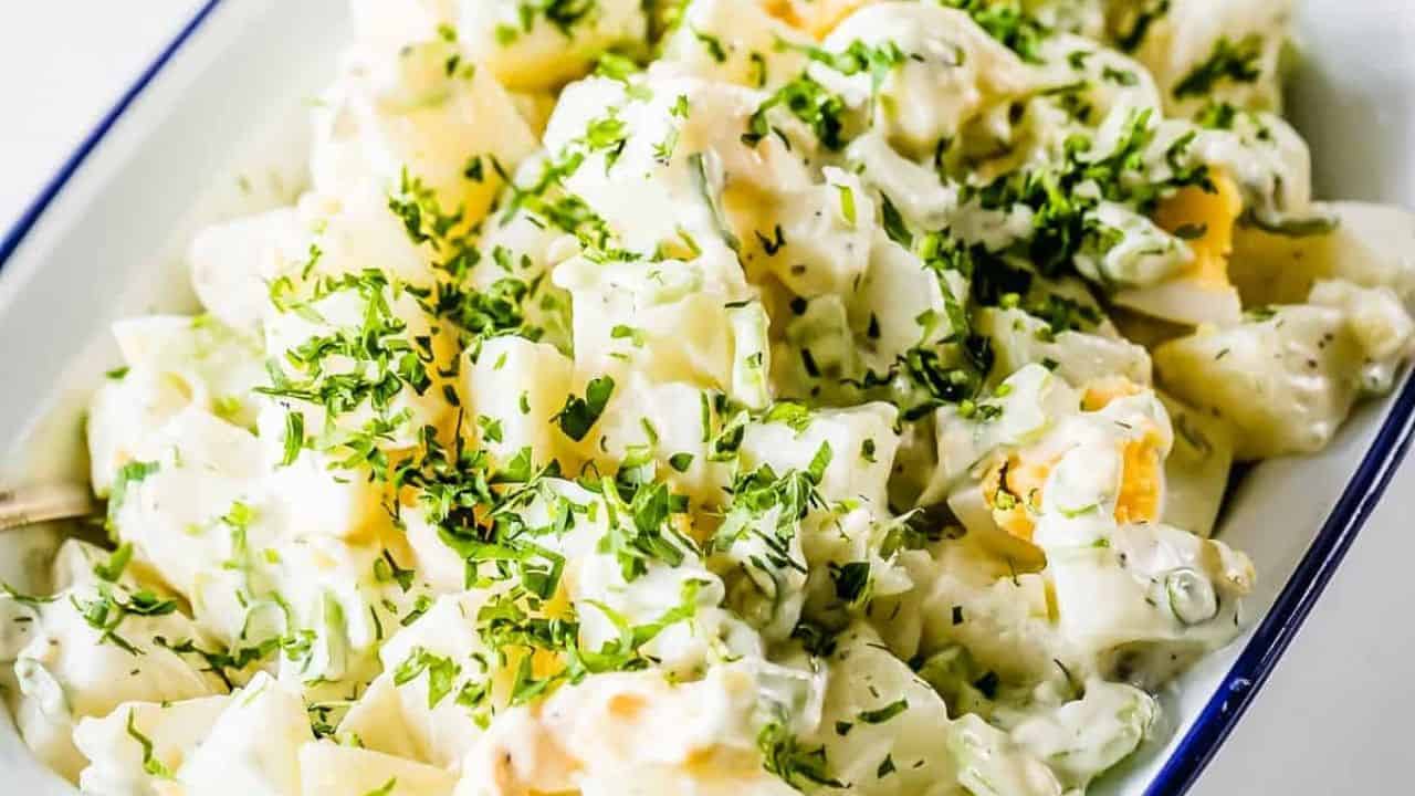 A close-up of creamy potato salad garnished with chopped parsley, served in a white dish with a blue rim.