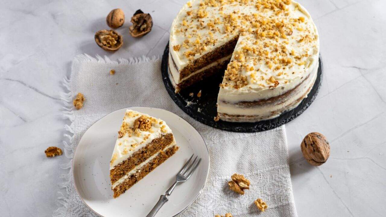 A slice of carrot cake with cream cheese frosting on a plate, garnished with walnuts. The whole cake is in the background.