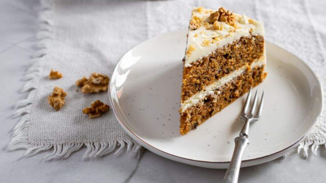 A slice of carrot cake with cream cheese frosting on a plate, garnished with walnuts. The whole cake is in the background.