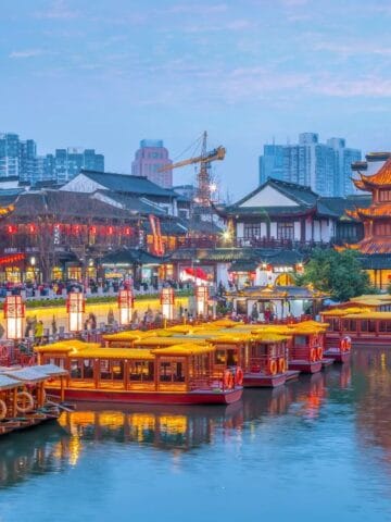 Traditional Chinese buildings and illuminated boats line a river at dusk, with modern city buildings visible in the background.