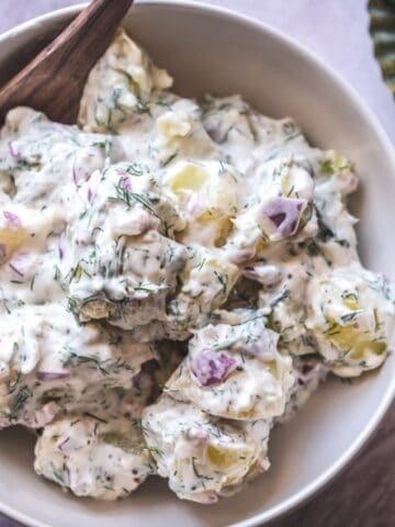 A bowl of creamy potato salad with chopped red onions and fresh dill, served with a wooden spoon; dill garnish is visible on a small plate nearby.