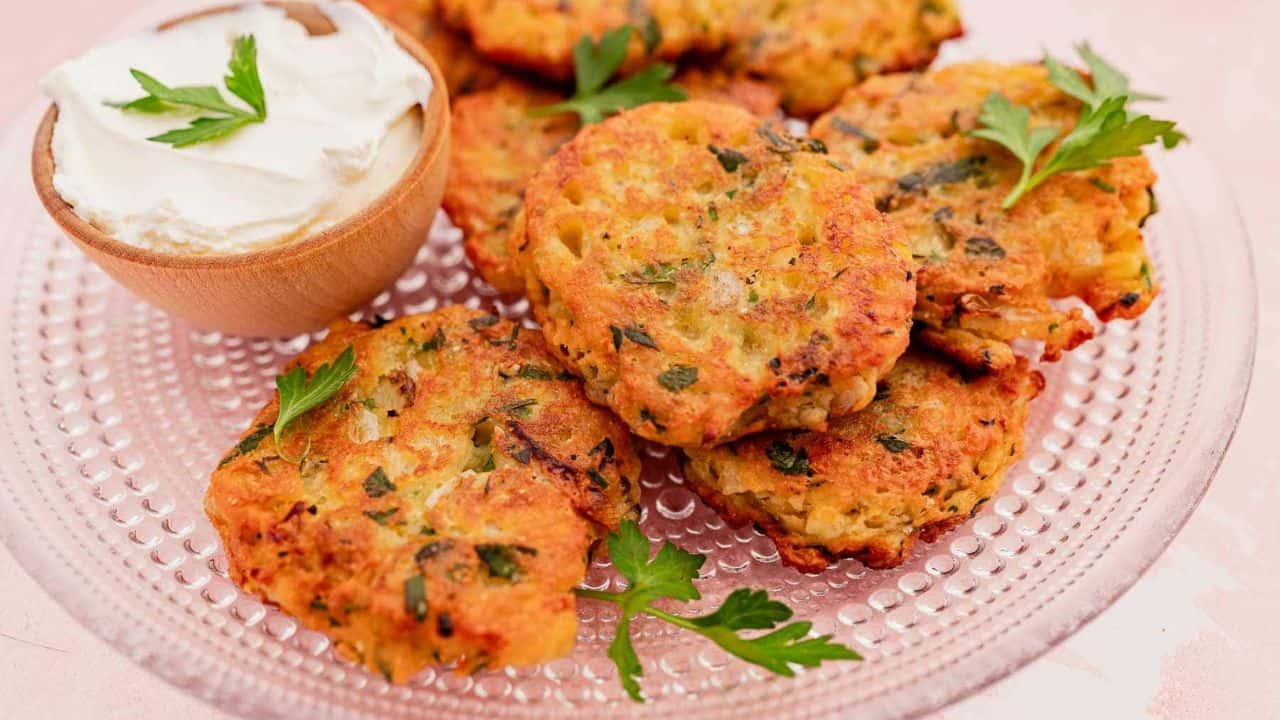 Five golden-brown vegetable fritters garnished with parsley are arranged on a glass plate next to a small bowl of sour cream.