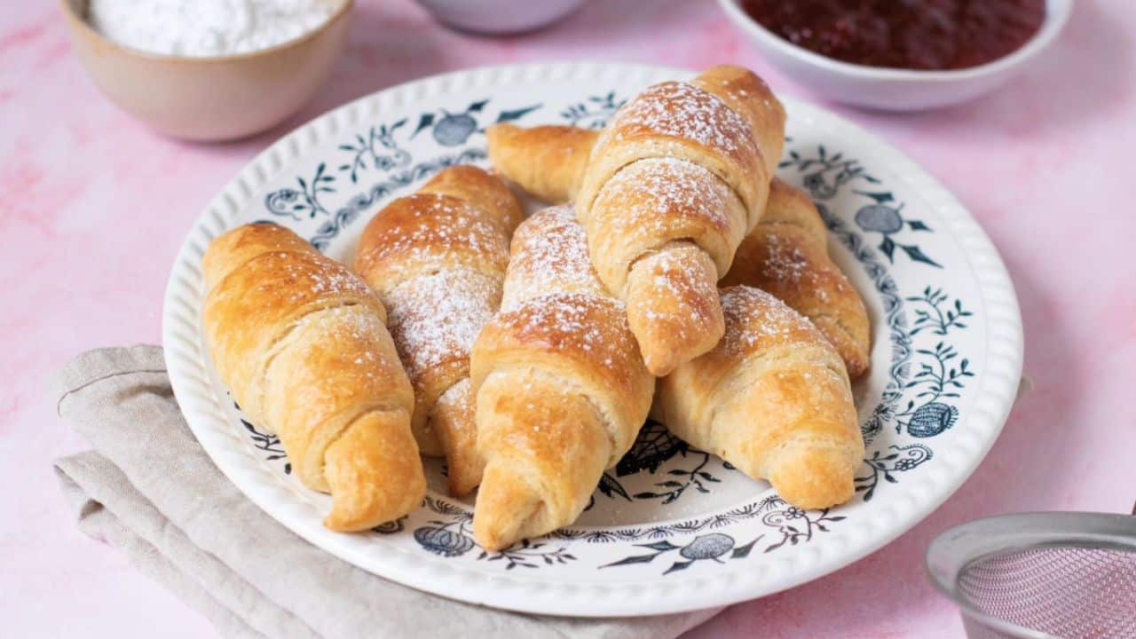 A plate of golden-brown croissants is set on a patterned dish. Nearby are bowls containing butter, flour, and red jam, with a beige cloth napkin under the plate.