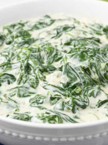 A white bowl filled with creamy spinach dip, with a wooden spoon partially submerged in the mixture, sits on a blue checkered cloth.