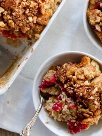 A serving dish and two bowls filled with fruit crumble topped with chopped nuts sit on a white cloth, with a metal spatula resting in the dish.