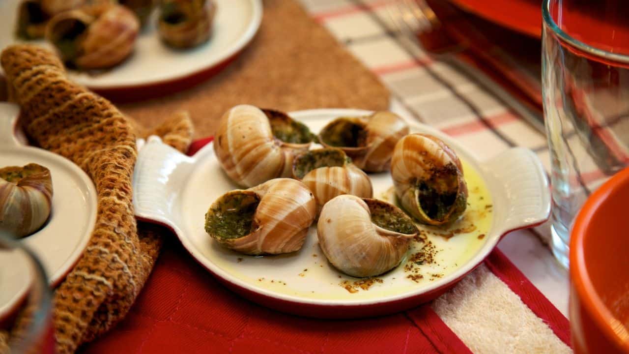A plate of cooked escargot with shells, filled with green herb butter sauce, is placed on a table set for a meal.