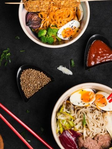 Three bowls of ramen with various toppings, including eggs, vegetables, and skewers, are arranged on a dark surface with sauces, seeds, and mushrooms scattered around.