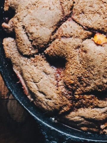 A close-up of a baked cobbler in a cast iron skillet with a wooden spoon, next to a bowl containing a serving of the cobbler with visible fruit pieces.