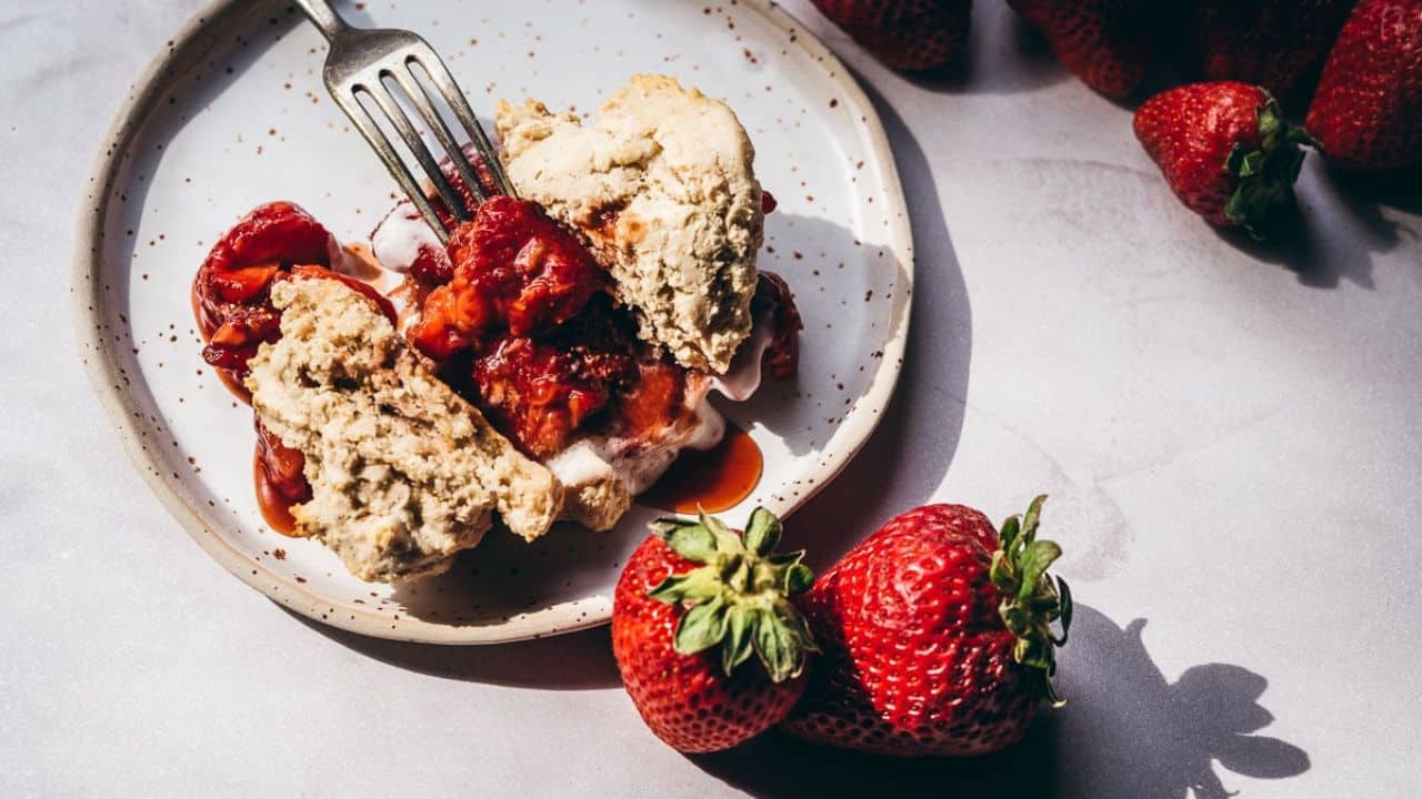 A plate with a biscuit split in half, topped with strawberries and cream, with a fork and fresh strawberries on the side.