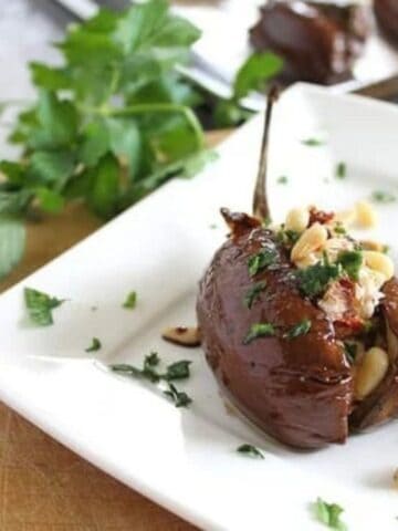 Two roasted eggplants stuffed with cheese, pine nuts, and herbs are served on a white plate, garnished with fresh parsley, with a fork and knife nearby.