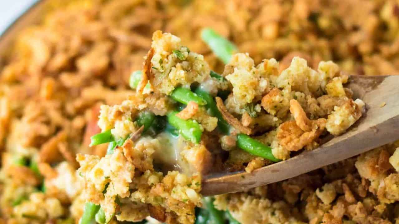 A close-up of a wooden spoon holding a serving of green bean casserole topped with crispy fried onions and breadcrumbs.