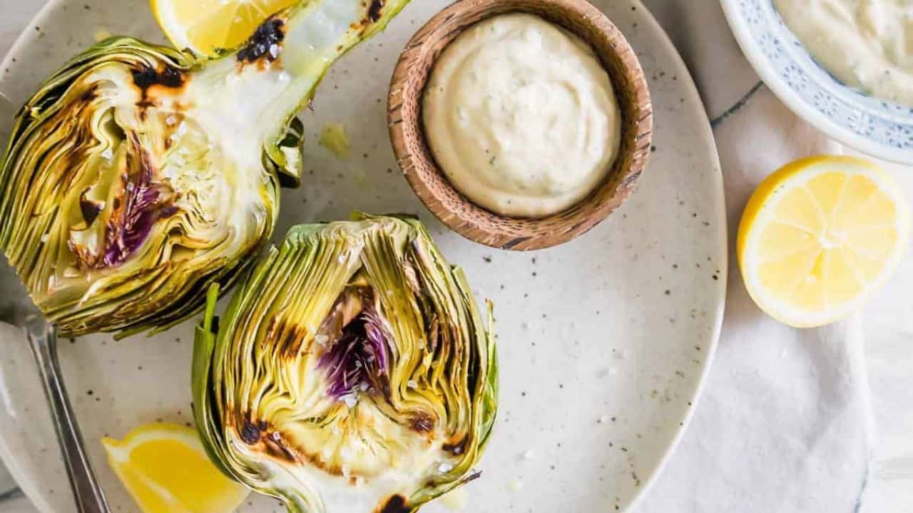 Grilled artichokes on a plate with dipping sauce and lemon.