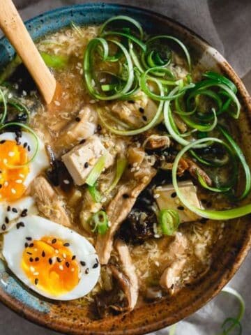 A bowl of ramen with sliced mushrooms, tofu, green onions, and halved soft-boiled eggs garnished with black sesame seeds; a wooden spoon rests inside the bowl.