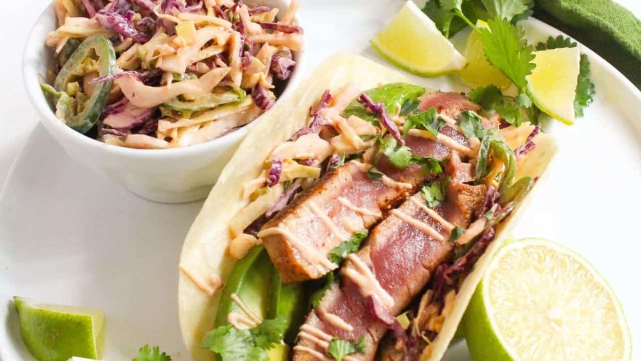 A plate with a seared tuna taco topped with slaw and cilantro, garnished with lime wedges, and a small bowl of slaw on the side.