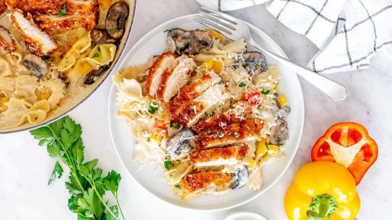 A plate of creamy bowtie pasta with mushrooms, sliced breaded chicken, grated cheese, and herbs, next to a skillet of pasta, fresh parsley, a yellow bell pepper, and a red bell pepper.