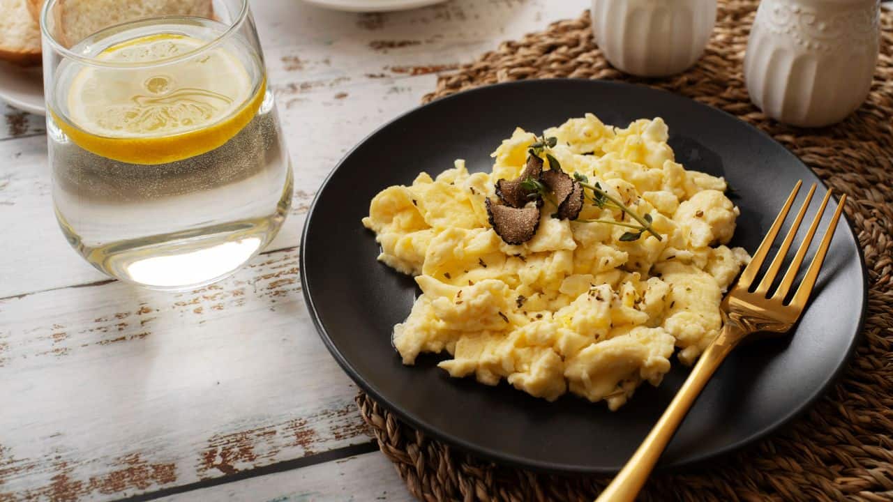 A plate of scrambled eggs garnished with herbs and truffle slices, served with a gold fork and a glass of water with a lemon slice.