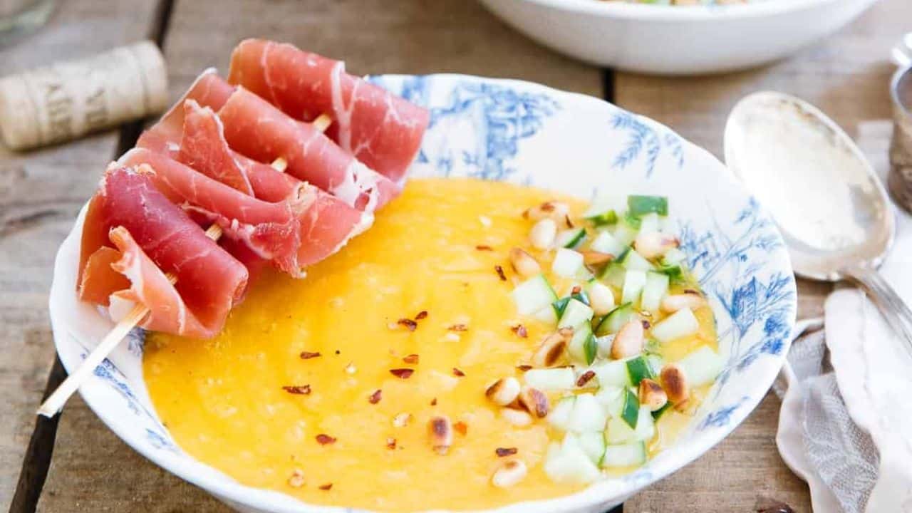 Mango melon soup with pickled cucumber and prosciutto skewer in a bowl.