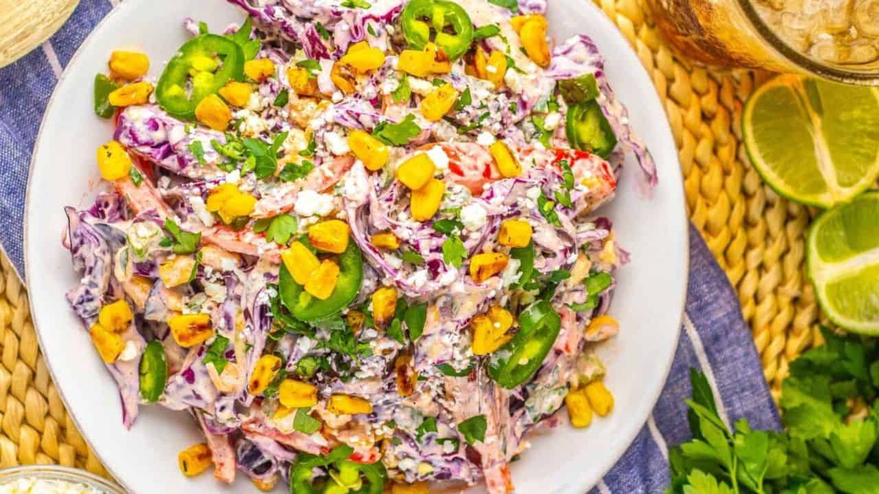 A plate of colorful salad with shredded cabbage, sliced jalapeños, roasted corn, chopped herbs, and crumbled cheese on a white plate, next to lime wedges and a drink.