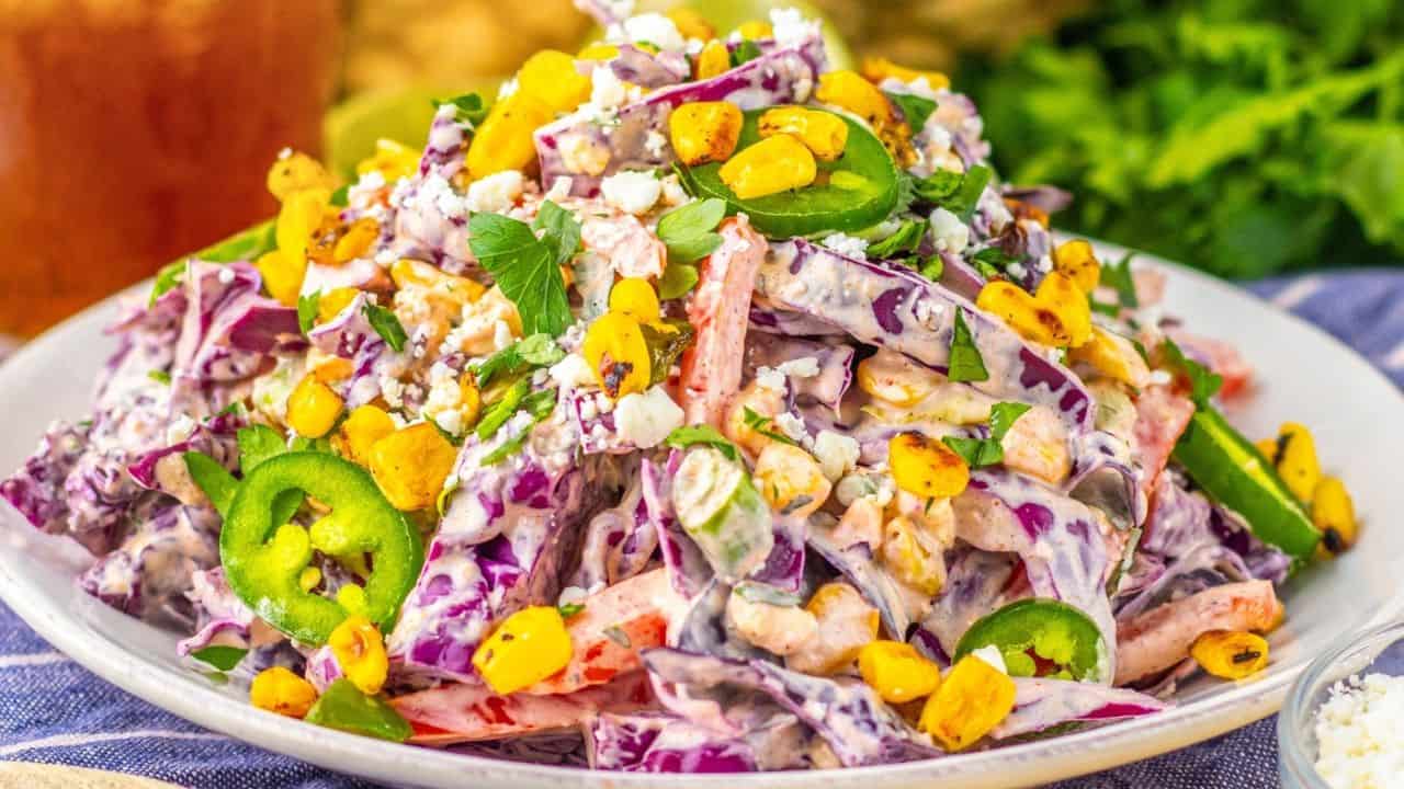 A plate of creamy salad with shredded purple cabbage, corn, sliced jalapeños, crumbled cheese, and chopped herbs, served on a white dish.