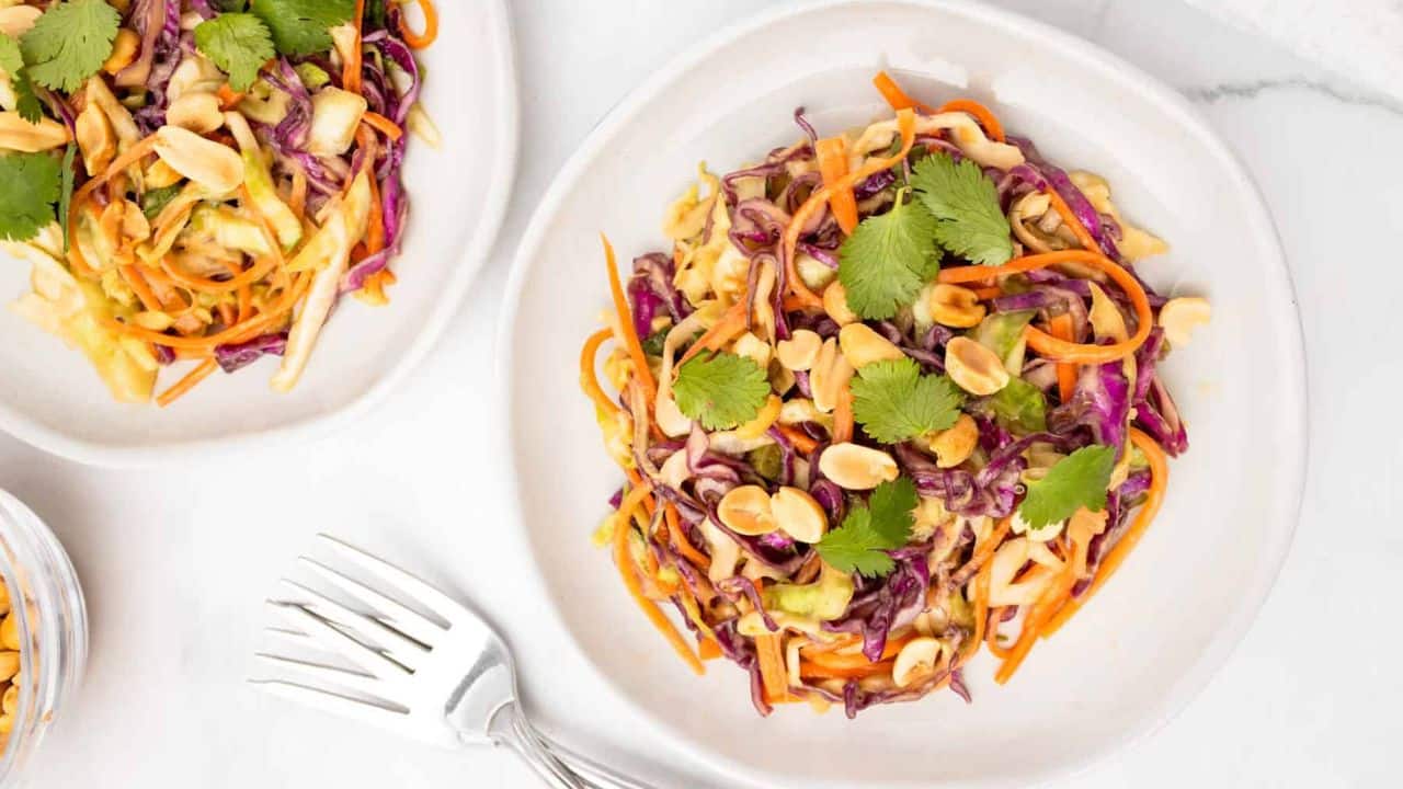 A plate of colorful cabbage and carrot slaw topped with peanuts and cilantro, with two forks and another serving on a partial plate nearby.