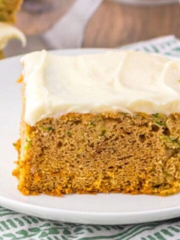 A slice of moist zucchini cake topped with creamy frosting is served on a white plate atop a green and white patterned napkin.