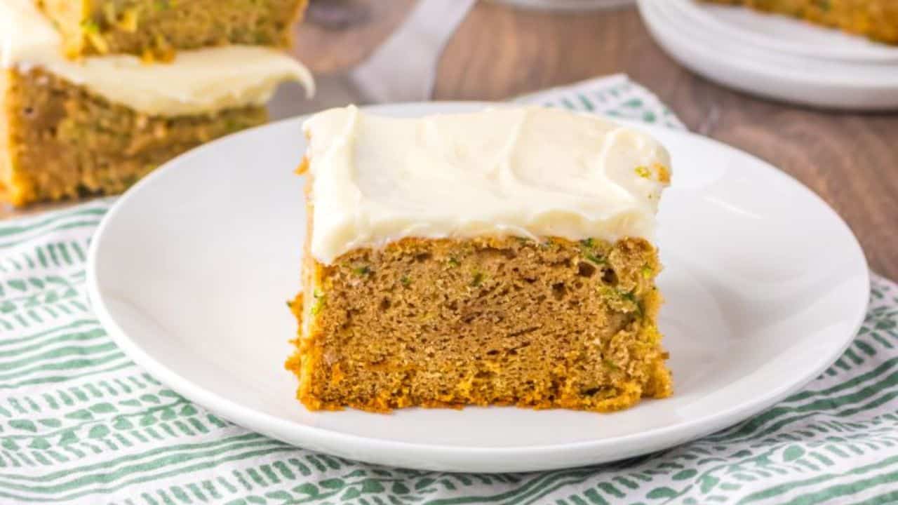 A piece of zucchini cake with white frosting on a white plate, sitting on a striped cloth with more cake in the background.