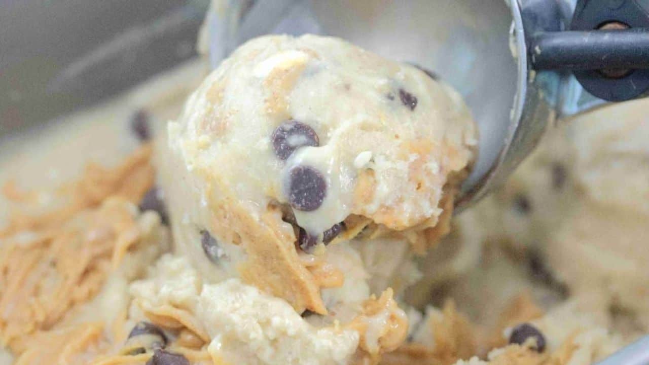 Peanut butter chocolate chip protein ice cream in a baking loaf pan with a spoon in it and an ice cream scoop and chocolate chips around it.