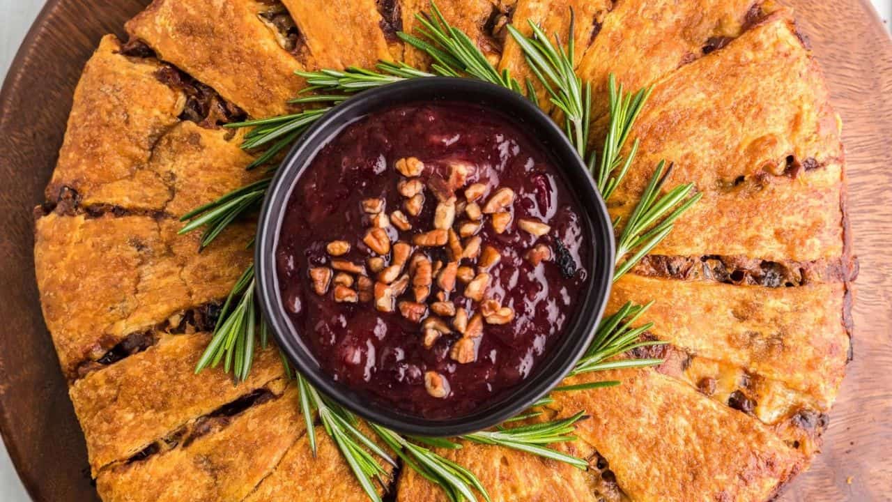 A round baked bread ring with a bowl of dark red dip topped with pecans in the center, garnished with fresh rosemary sprigs.