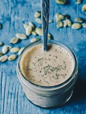 A glass jar filled with a creamy beverage, topped with herbs, sits on a blue wooden surface with a spoon inside and green cardamom pods scattered around.