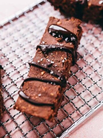 Two rectangular chocolate brownies with chocolate drizzle rest on a metal cooling rack, with more brownies visible in the background.