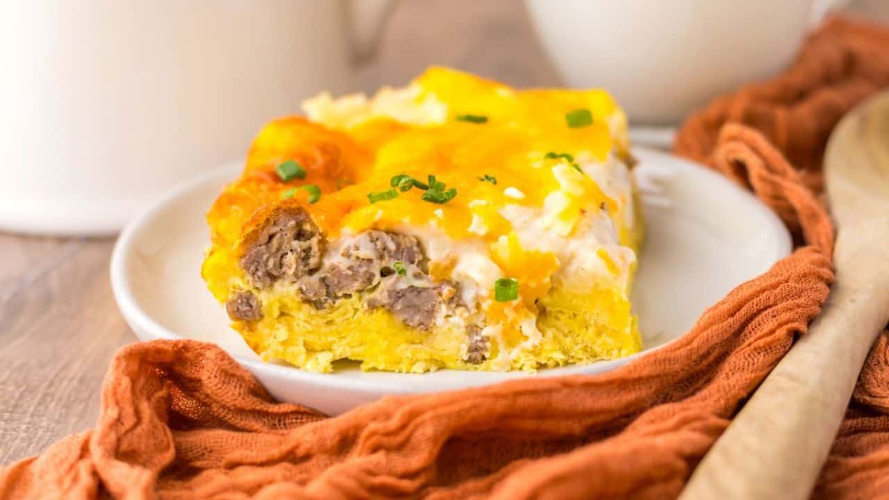 A slice of breakfast casserole with sausage, eggs, and cheese is served on a white plate beside an orange cloth and a wooden spoon.