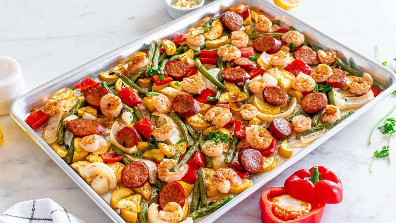 A sheet pan filled with cooked shrimp, sliced sausage, bell peppers, onions, and zucchini on a white surface.