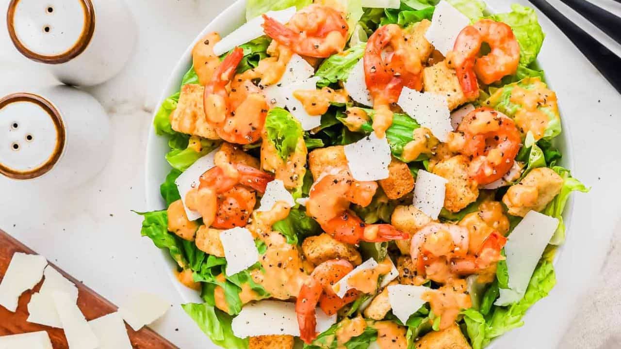 A fresh shrimp Caesar salad with romaine lettuce, shrimp, croutons, shaved parmesan cheese, and Caesar dressing is served in a white bowl with black utensils on a marble table.