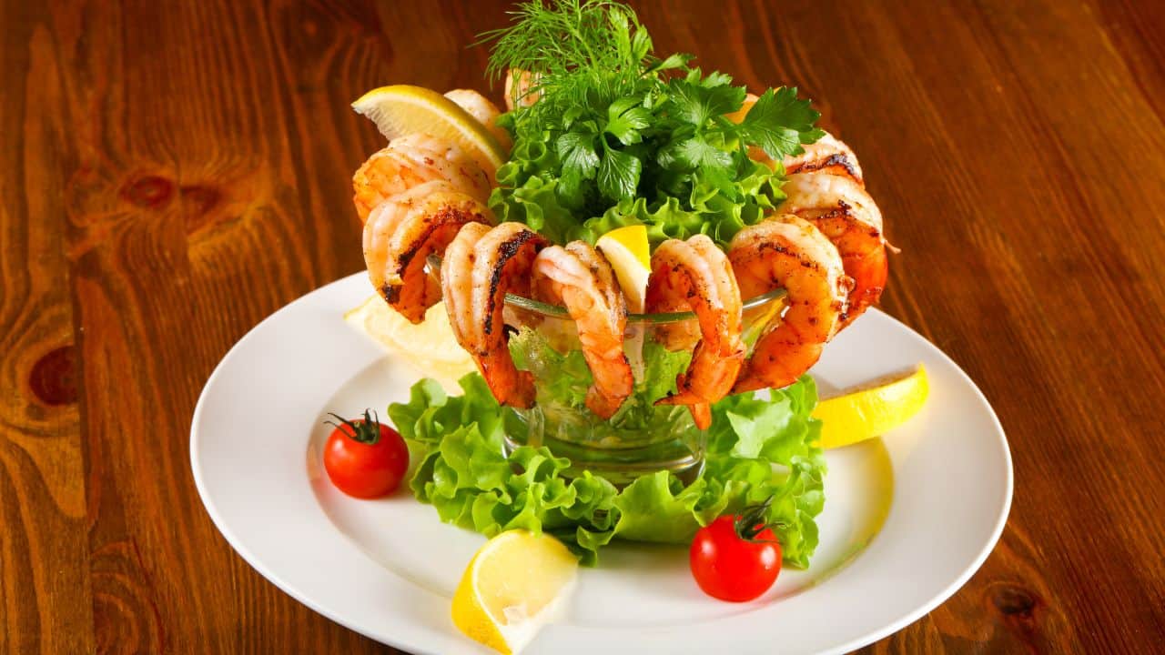 A shrimp cocktail served in a glass with lettuce, garnished with lemon wedges, cherry tomatoes, and fresh herbs on a white plate.