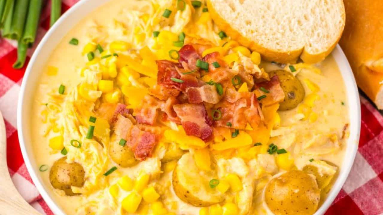 A bowl of creamy potato corn chowder topped with bacon and cheese, next to a wooden spoon, bread slices, and a green plant on a red checkered cloth.