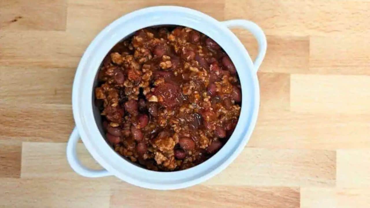 Image shows close-up of a spoonful of chili with beans, cheese, and corn, held above a pot filled with the same dish, placed on a wooden surface.
