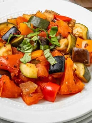 A white plate with a serving of ratatouille, featuring chopped tomatoes, zucchini, eggplant, bell peppers, and garnished with fresh basil.