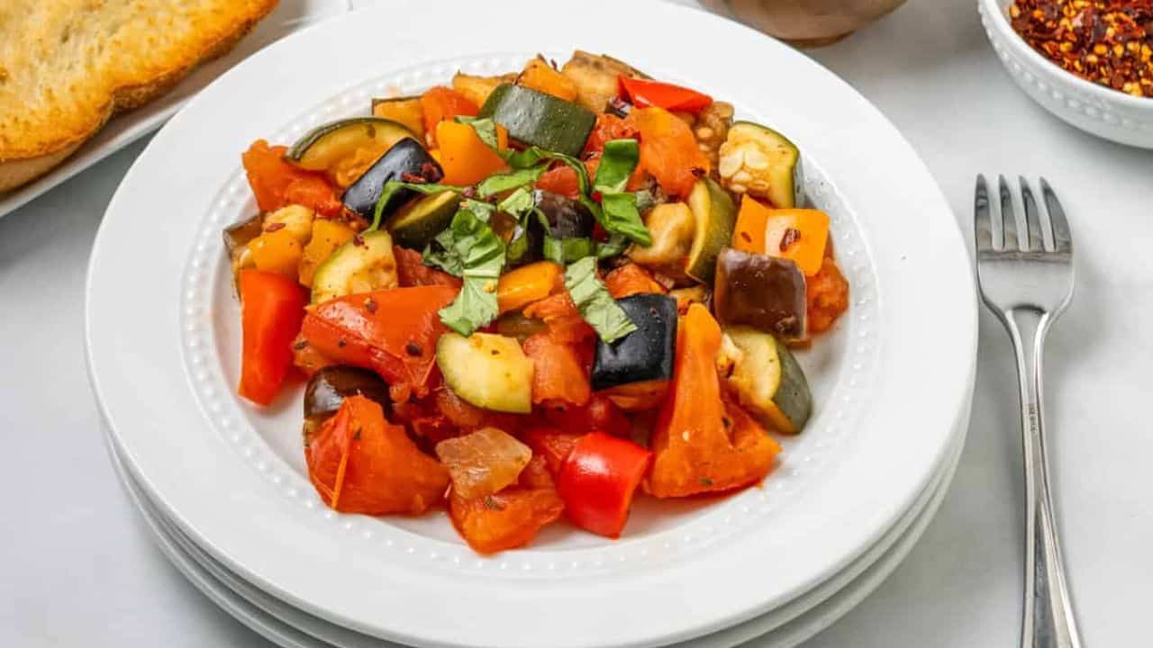 A white plate with a serving of ratatouille, featuring chopped tomatoes, zucchini, eggplant, bell peppers, and garnished with fresh basil.
