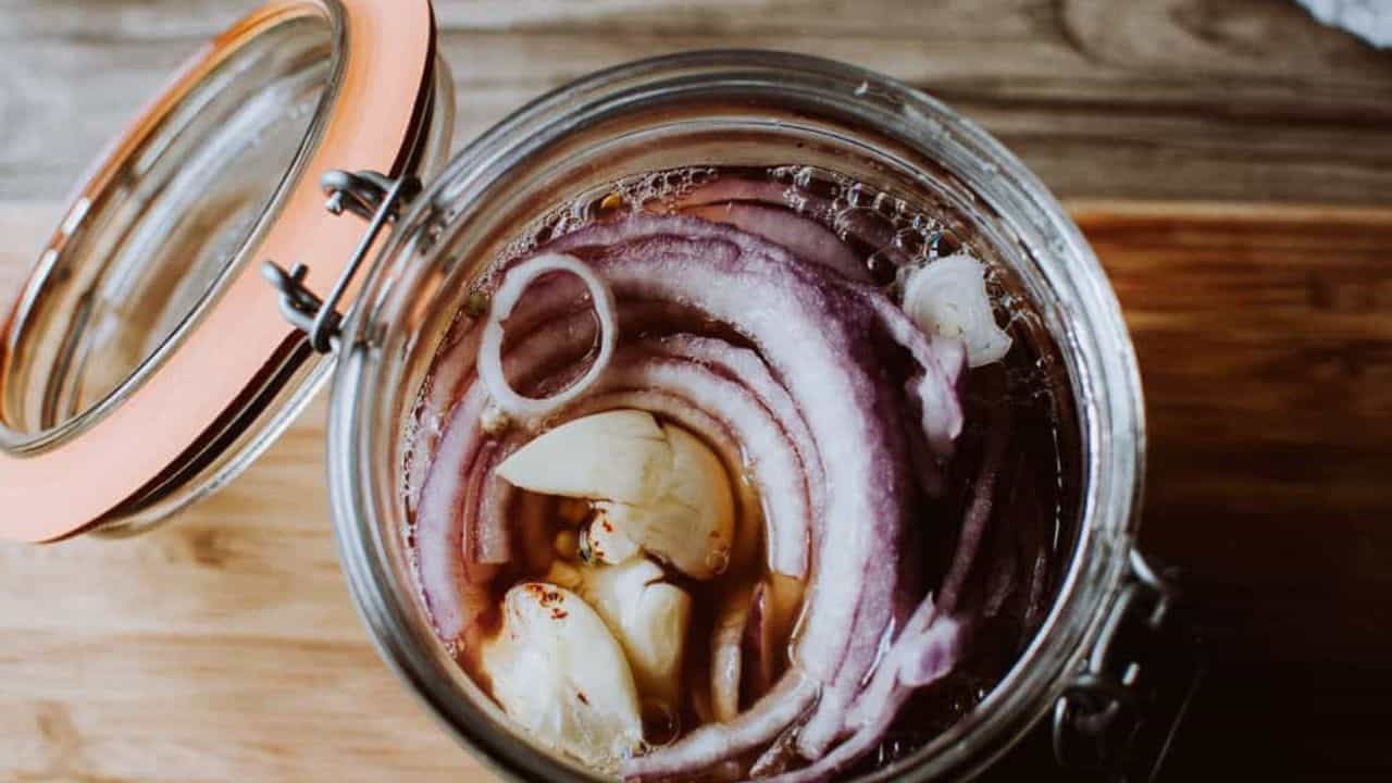 An open glass jar containing sliced red onions and garlic cloves in liquid, placed on a wooden surface.