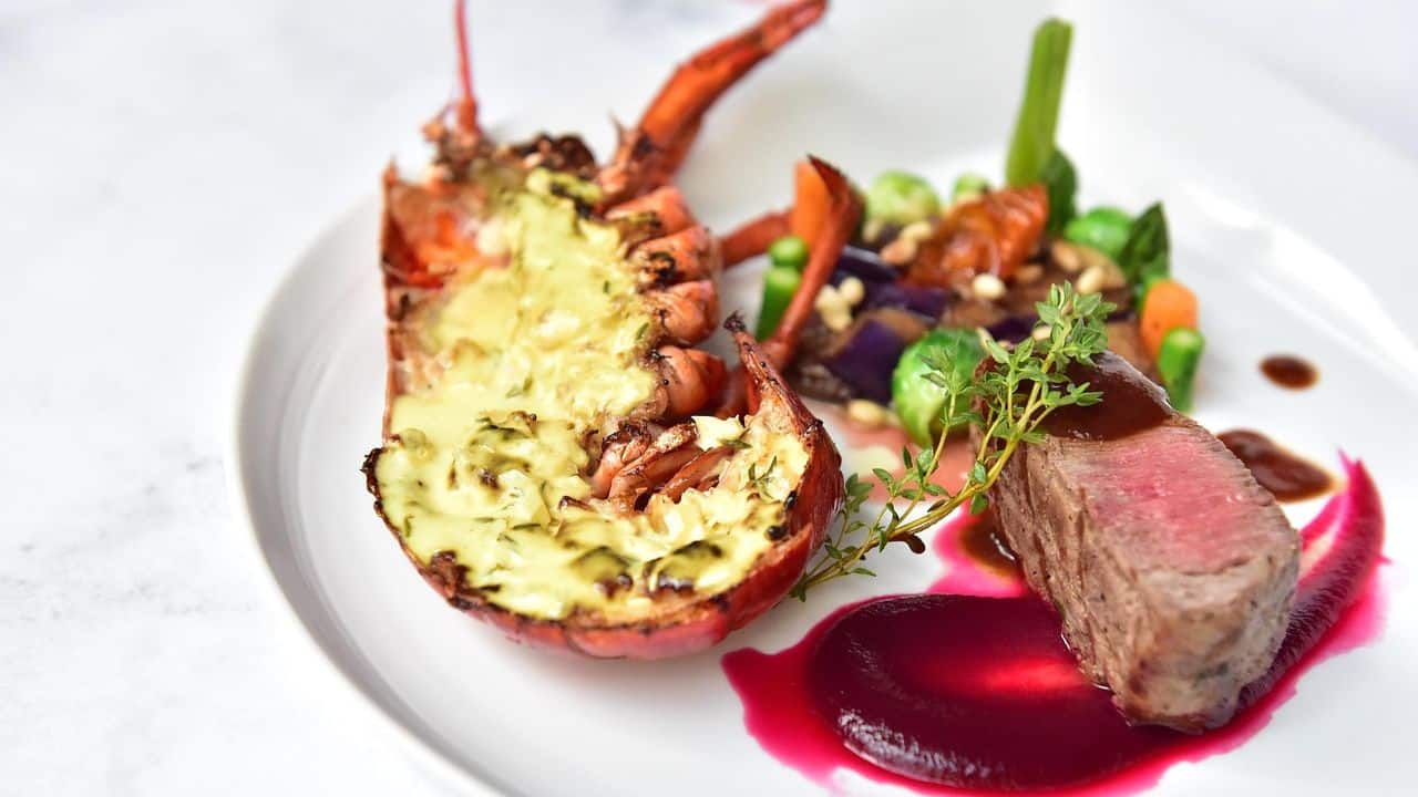 A plated dish featuring half a grilled lobster, a piece of beef steak, assorted vegetables, and a beet puree garnish on a white plate.